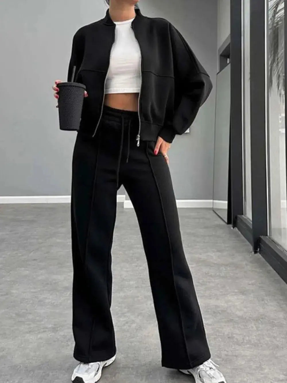 Woman wearing black baseball collar zip up outerwear and matching drawstring pants set with white crop top and sneakers indoors