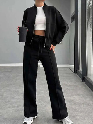 Woman wearing black baseball collar zip up outerwear and matching drawstring pants set with white crop top and sneakers indoors