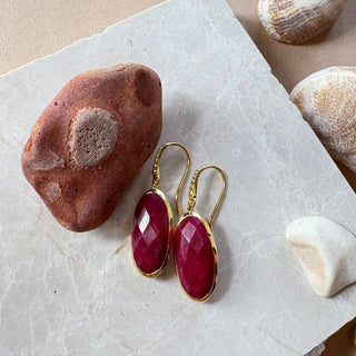 Charis Oval Gemstone Earrings handcrafted with ruby and 22K gold on recycled brass displayed on marble surface with stones.