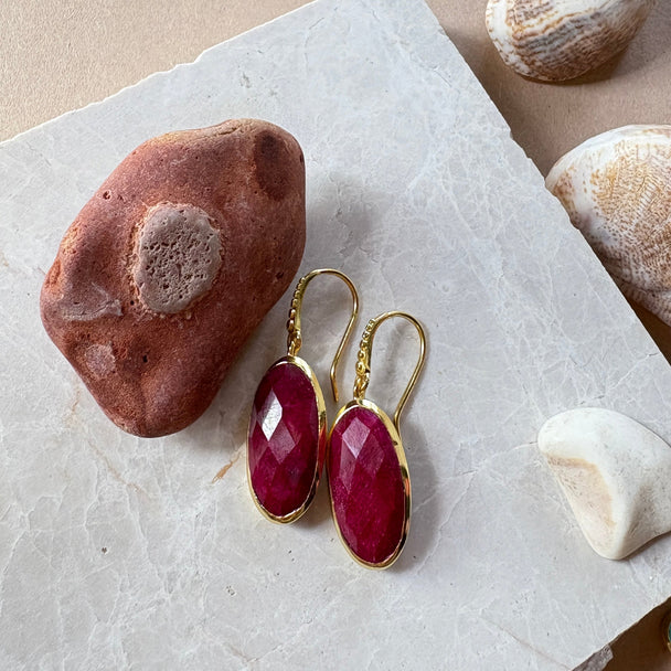 Charis Oval Gemstone Earrings handcrafted with ruby and 22K gold on recycled brass displayed on marble surface with stones.