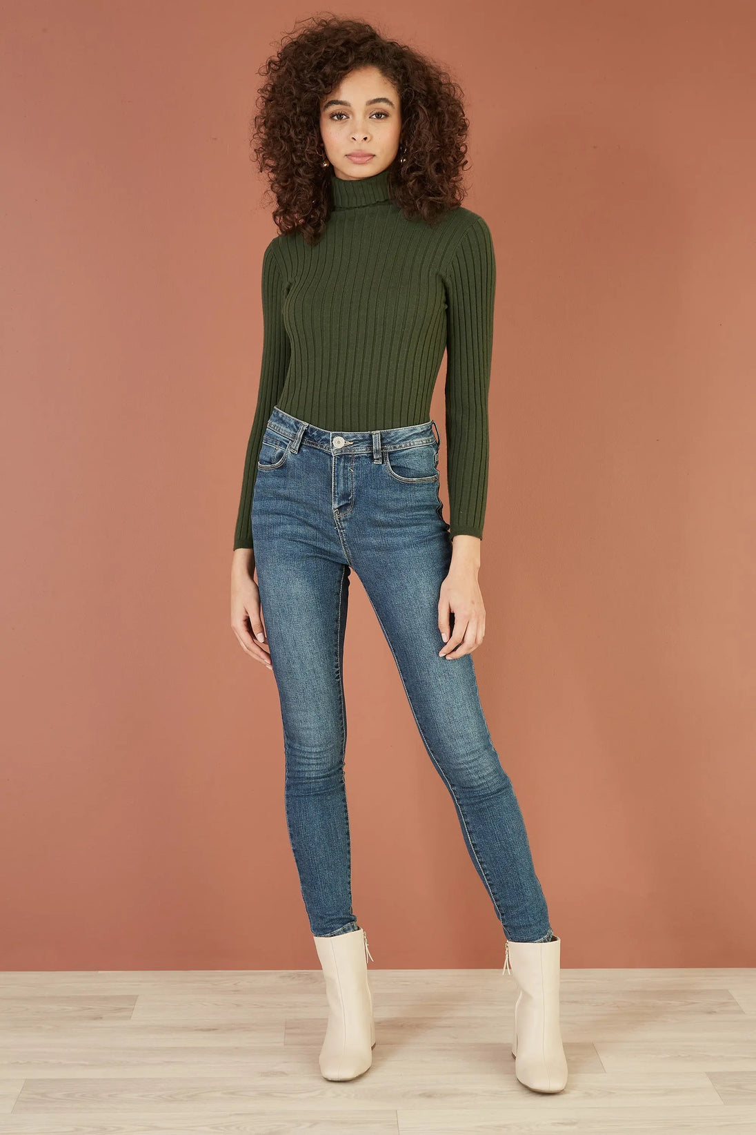 Woman wearing Yumi green ribbed knit roll neck jumper with blue jeans and beige boots against brown background