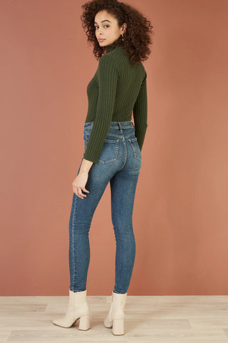 Woman wearing Yumi green ribbed knit roll neck jumper with fitted silhouette and blue skinny jeans.