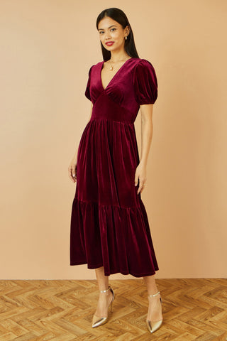 Woman wearing Yumi Burgundy Velvet Midi Dress with V neckline and tiered skirt against beige background