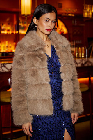 Woman wearing Yumi Coffee Luxe faux fur coat with oversize collar over blue sequin dress in warm bar setting