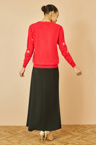 Woman wearing red gold foil star print relaxed fit jumper with black maxi skirt and gold heels, shown from back
