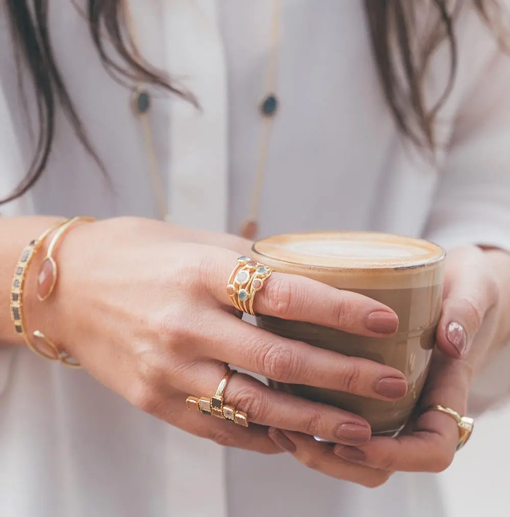 Close-up of a hand wearing the Iona Entwined Ring with green onyx, amethyst, moonstone, and labradorite gemstones, holding a cup of latte.