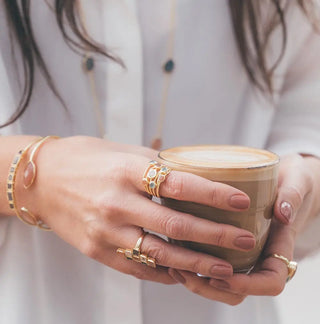 Close-up of a hand wearing the Iona Entwined Ring with green onyx, amethyst, moonstone, and labradorite gemstones, holding a cup of latte.